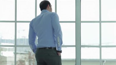 Kendine güvenen mavi gömlekli bir işadamı pencerenin kenarında durup, düşünceli bir ifadeyle saatine bakıyor. Yumuşak doğal ışığın içeri aktığı parlak ofis ortamı, hırs ve başarı atmosferini yükseltiyor.