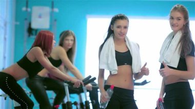 Bir grup güçlü bayan sporcu canlı bir spor salonunda güven içinde poz veriyor, spor yaparken bir yandan da sporcuları destekliyorlar. Arkadaşları aktif olarak durağan bisikletlerle bisiklet sürmekle meşguller.