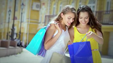 Biri parlak sarı elbiseli, diğeri beyaz elbiseli iki neşeli kız, canlı bir açık hava alışveriş merkezinde dolaşırken renkli alışveriş torbalarını zevkle incelerler. Işıl ışıl gülüşleri ve kaygısız tavırları...