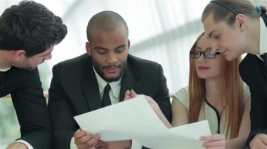 Aralarında kızıl saçlı bir kadının da bulunduğu dört kendine güvenen işadamı, gösterişli bir ofis masasında belgeleri tartışırken görülüyor. Gülüşleri ve odaklanmış ifadeleri bir işbirliği ve başarı ortamı yaratıyor.