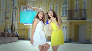 İki neşeli kız, biri beyaz elbiseli, diğeri de güneşli sarı elbiseli, ellerinde renkli alışveriş torbalarıyla neşe içinde poz veriyorlar. Işıl ışıl gülüşleri ve neşeli jestleri arkadaşlığın ve kaygısız alışveriş maceralarının özünü yakalar.