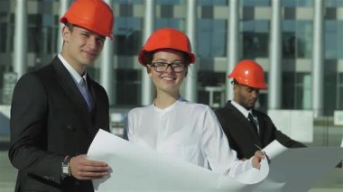 Üç kendine güvenen mimar, parlak turuncu kasklar takıyor, olay yerinde duruyor, şevkle planları gözden geçiriyorlar. Ekip çalışmaları ve profesyonellikleri, bu heyecan verici inşaat projesinin geleceğini öngördüklerinde parlıyor.