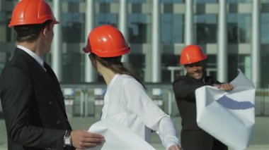 Parlak turuncu miğferler takmış üç kendine güvenen mimar hareketli bir inşaat sahasında duruyor ve şevkle planları inceliyor. Dinamik takım çalışmaları ve profesyonel tavırları yenilikçi yapı fikirlerine olan bağlılıklarını yansıtıyor.