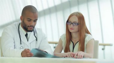 Gıcır gıcır beyaz önlük giymiş odaklanmış bir doktor göğüs röntgenini incelerken bir hastayla detaylı bir tartışmaya girer. Parlak, modern klinik kurulumu profesyonellik ve bakım atmosferini artırarak,