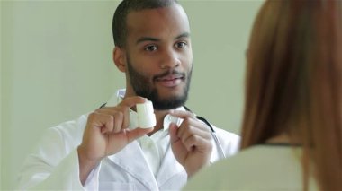 Şefkatli bir doktor, sıcak bir gülümsemeyle, parlak, modern bir klinikteki bir hastaya reçeteli ilacı dikkatlice açıklar. Bu etkileşim hasta bakımının önemini vurguluyor, çünkü doktor ilaç şişesini tutuyor,