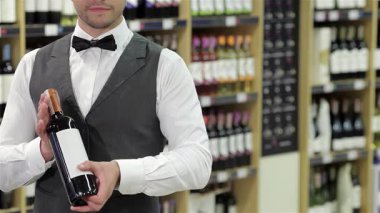 Şık bir smokin giymiş kendine güvenen bir erkek şarap garsonu, sofistike bir şarap dükkanında şarap şişesini gururla tutar. Etkileyici bir dizi şarap şişesi ile çevrelenmiş, uzmanlık ve zarafeti somutlaştırıyor, şarap meraklılarını şımartmaya davet ediyor.