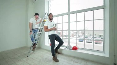 İki hevesli genç inşaatçı, biri merdiven üzerine tünemiş ve diğeri de büyük bir pencerenin yanında, ellerinde boya kutuları ve parlayan başparmaklar olan ferah bir dairede güven içinde duruyorlar. Gülüşleri pozitiflik ve takım çalışması yayar, ruhu somutlaştırır.