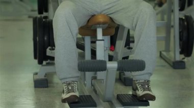 Konforlu egzersiz kıyafetleri giymiş odaklanmış genç bir adam göğüs kaslarını ağırlık sırasının üzerinde yoğun bir şekilde çalıştırıyor. Kararlılık ve güç gösterisi yapıyor. Spor salonu enerji ile uğulduyor, çeşitli spor aletleri ve ağırlıklarla.