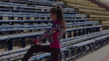 Odaklanmış genç bir kız dinamik bir esneme egzersizi yapıyor, bacağını canlı bir atletik kıyafetle boş stadyum tribünlerinin arka planına kaldırıyor. Bu enerjik sahne hazırlık ve kararlılığın ruhunu yansıtıyor.