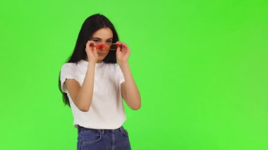 Uzun, dalgalı siyah saçlı çarpıcı genç bir kadın, modaya uygun pembe güneş gözlüklerini canlı yeşil bir arka plana göre ayarlıyor. Onun gündelik beyaz tişörtü ve rahat tavırları neşeli ama şık bir hava yayıyor, izleyicileri kendi tarzlarını benimsemeye çağırıyor.