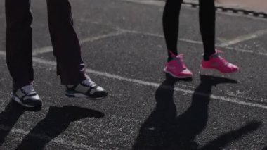 Canlı atletizm kıyafetleri giymiş iki genç kız, güneş ışığında enerjik bacak ısıtma egzersizleri yapıyor, bağlılıklarını ve güçlerini sergiliyorlar. Sahne, gençlik kararlılığının ve rekabetin heyecanını yansıtıyor.