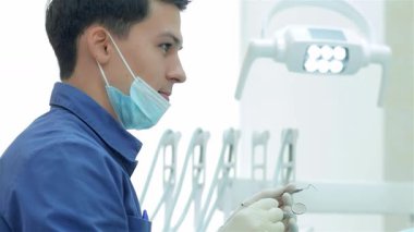 Mavi üniforma ve koruyucu maske takan kendini işine adamış bir dişçi asistanına bakarken dişçi aletini ustalıkla inceliyor. Parlak, klinik ortam bu diş muayenesinin profesyonelliğini artırıyor.