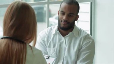 Neşeli bir Afrikalı adam parlak bir klinikte düşünceli bir kadın doktorla görüşüyor. Doktor dikkatle dinliyor, elinde bir pano var, reçete yazmaya hazırlanıyor, profesyonelliği ve şefkati somutlaştırıyor.