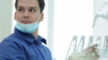 Mavi üniformalı neşeli bir dişçi, parlak ve modern bir klinikte dişçi aletini tutarak asistanına sıcak bir şekilde gülümsüyor. Atmosfer pozitiflik ve profesyonellikle dolu. Hasta bakımında işbirlikçi bir ruh sergiliyor.