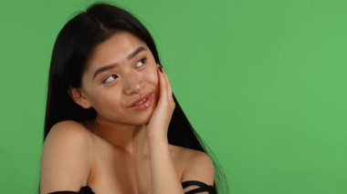 Çekici genç bir Asyalı kadın canlı yeşil renkli bir arka plana karşı rüyasında poz veriyor, neşeli gülüşü sıcaklığı ve kendine güveni yayıyor. Uzun, dalgalı saçları ve narin özellikleriyle güzelliği ve canlılığı somutlaştırarak izleyicileri paylaşmaya davet ediyor.