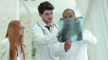Modern bir hastane ortamında, gözlüklü bir kadın ve beyaz önlüklü iki adam da dahil olmak üzere üç kendine güvenen doktor, sağlıklı akciğerlerin röntgenini yakından inceliyor. Onların odaklanmış ifadeleri ve takım çalışması tıbbi uzmanlığın önemini vurgular