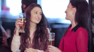 İki neşeli kız kadehlerini kaldırarak, kahkaha ve arkadaşlıkla dolu canlı bir atmosferle çevrelenmiş bir şekilde kadeh kaldırıyorlar. Bar ışıklarının sıcacık parıltısı, içkinin keyfini çıkarırken ve arkadaşlarıyla neşeli anları paylaşırken neşeli havayı arttırır.
