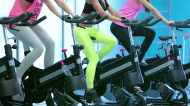 Canlı bir fitness sınıfı, hareketsiz bisikletler üzerinde pedal çeviren atletik kadınları sergiliyor. Güç ve dostluk saçıyor. Renkli spor kıyafetleri ve odaklanmış ifadeler ilham verici bir atmosfer yaratıyor.