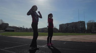 İki genç kız, parlak bir gökyüzüne karşı siluetlenmiş, bir pistte enerjik ısınma egzersizleri yapıyorlar, vücutlarını yaklaşan yarışa hazırlıyorlar. Atletik formları güç ve kararlılık yayar, gençliğin güzelliğini ve uygunluğunu gözler önüne serer.