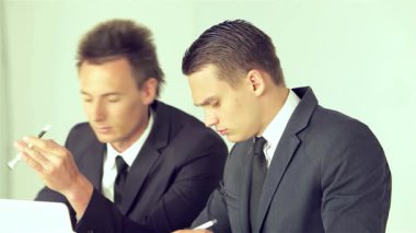 İki keskin giyimli iş adamı, biri şık saç kesimi olan, düzgün bir dizüstü bilgisayardaki verileri analiz ederek, odaklanmış bir tartışmaya giriyorlar. Ciddi ifadeleri ve özenli vücut dilleri yüksek riskli bir kurumsal ortamı yansıtıyor.