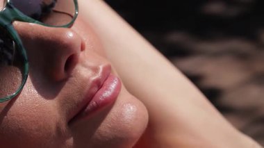 Göz kamaştırıcı bir yakın plan, güneşte güneşlenen neşeli genç bir kadını, bronzlaşmış tenini aydınlatan parlak gülüşünü yakalar. Güneş gözlükleriyle parlak güneş ışığını yansıtıyor. Yaz rahatlığının ve kaygısızlığın özünü somutlaştırıyor.