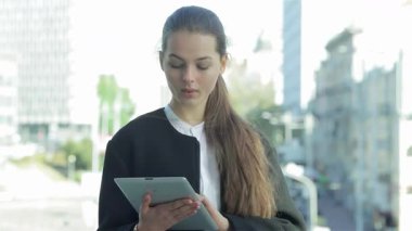 Dengeli bir iş kadını dışarıda duruyor, tabletindeki partnerleriyle özenle iç içe geçiyor, profesyonellik ve güven yayıyor. Parlak kentsel zemin, başarı ve işbirliği atmosferini güçlendiriyor ve onu önemli bir oyuncu olarak gösteriyor.