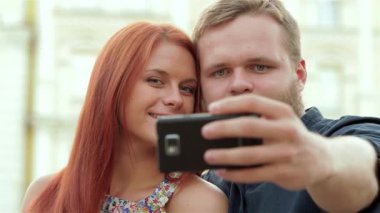 Neşeli genç bir çift, uzun kızıl saçlı bir kadın ve şık sakallı bir adam, şehir bankında otururken mutlu bir şekilde akıllı telefonlarıyla selfie çekiyorlar. Güneş ışıl ışıl parlıyor, neşeli bir şehre karşı gülüşlerini aydınlatıyor.