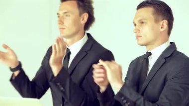 İki keskin giyimli iş adamı, biri dağınık saçlı, diğeri parlak saçlı, canlı bir iş konferansında coşkulu bir şekilde bir konuşmacıyı alkışlıyor. İfadeleri pozitiflik ve bağlılık yayıyor, profesyonel bir atmosfer sergiliyor.