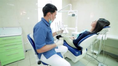 Kıtır üniforma giymiş yetenekli genç bir dişçi dişçi dişçi sandalyesindeki hastasına doğru eğiliyor ve ona dostça bir gülümseme takınıyor. Diş aletleri ışıldarken, parlak, modern klinik kurulumu bakım ve profesyonellik atmosferini artırıyor