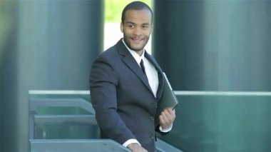 Başarılı bir Afrikalı işadamı elinde bir tablet tutarken gururla dışarıda duruyor, güven ve neşe saçıyor. Resmi kıyafeti ve ışıl ışıl gülüşü profesyonel yolculuğunun zaferini yansıtıyor. Yeni fırsatlar ve ortaklıklar yaratıyor.