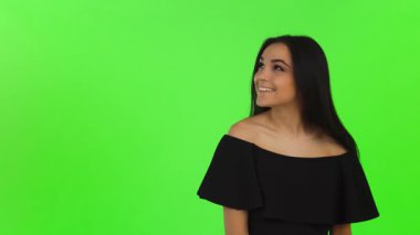 Şık siyah elbiseli çarpıcı genç bir kadın neşe saçıyor. Işıl ışıl gülümseyip, parlak yeşil renkli arka planı işaret ediyor. Bu büyüleyici stüdyo portresi özgüven ve zarafet yayıyor. Reklam ve promosyon için mükemmel.