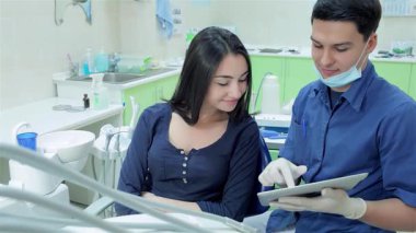 Mavi üniformalı neşeli bir dişçi dişçi, dişçi koltuğunda oturan genç bir kıza tablette x-ray 'ini gösterirken sıcak bir şekilde gülümsüyor. Parlak klinik ortamı pozitif enerji yayıyor, diş muayenelerini dostça ve ulaşılabilir kılıyor.