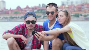 Üç genç arkadaş nehir kenarında oturuyor, gülüyor ve birbirlerinin resimlerini çekiyorlar, unutulamaz anılar yaratıyorlar. Canlı yaz atmosferi onların neşeli ifadelerini artırıyor, dostluğun ve eğlencenin özünü gözler önüne seriyor.