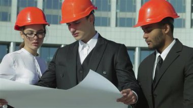 Canlı turuncu miğferli üç kendine güvenen mimar hareketli bir inşaat sahasında dikilip planlarını dikkatle gözden geçiriyorlar. Odaklanmış ifadeleri inovasyon ve takım çalışmasının ruhunu somutlaştıran kararlılık ve işbirliğini yansıtır.
