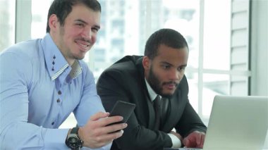 İki kendine güvenen iş adamı, biri sıradan giyinmiş, diğeri de şık takım elbiseli, dizüstü bilgisayarlarıyla şık bir ofis ortamında meşgul olurken gülümsüyorlar. Parlak, havadar ortam, profesyonelliğin bir karışımını sergileyerek takım çalışmalarını geliştirir.