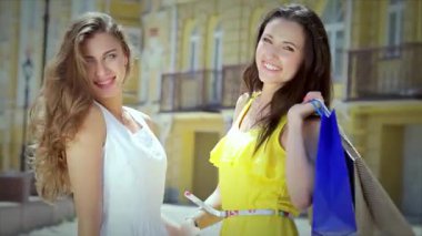 Biri beyaz elbiseli, diğeri parlak sarı elbiseli iki şık kız renkli alışveriş torbaları tutarken neşeli bir anı paylaşıyorlar. Işıl ışıl gülüşleri ve kaygısız tavırları arkadaşlığın özünü ve alışverişin heyecanını yansıtıyor.
