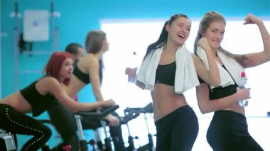 Canlı bir spor salonu ortamında, iki fit arkadaş kaslarını gururla sergiliyor, kendilerine güvenlerini ve arkadaşlıklarını sergiliyorlar. Bu arada, arkadaşları sabit motorlarla kardiyo yapıyor, motivasyon dolu enerjik bir atmosfer yaratıyorlar ve