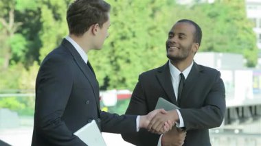 İki profesyonel iş adamı, biri açık tenli, diğeri koyu tenli, hareketli bir şehir arka planında sıcak bir şekilde el sıkışıyor, başarılı bir ortaklığı simgeliyor. Gülüşleri pozitiflik ve hırs saçıyor.