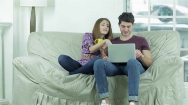 Neşeli genç bir çift rahat bir koltukta oturur, çay yudumlar ve bir dizüstü bilgisayarla uğraşırken kahkahalarını paylaşırlar. Neşeli ifadeleri sıcaklık ve mutluluk saçıyor, dostluk ve rahatlama dolu hoş bir atmosfer yaratıyor.