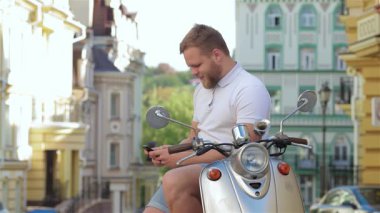 Sakallı neşeli bir genç adam şık scooterının üzerinde oturuyor, akıllı telefonuyla canlı bir sohbete dalmış. Canlı kentsel zemin, özgürlük ve macera hissini arttırıyor. Bu anı hem rahat hem de heyecan verici kılıyor.