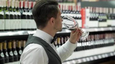 Şık bir yelek ve papyonlu şık bir genç adam bardaktan zarif bir şekilde yudumluyor. Etrafı şarap şişeleriyle çevrili, iyi stoklanmış bir içki dükkanında. Kendine güveni ve kaliteye olan keskin bakışı şarap yapımının sanatını yansıtıyor.