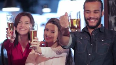 Aralarında iki kadın ve bir erkeğin de bulunduğu genç bir grup neşeyle bira bardaklarını kaldırıyor, mutluluk ve dostluk saçıyor. Barın hareketli atmosferi birlikte kutlama yaparken kahkahalar ve tezahüratlarla dolu.