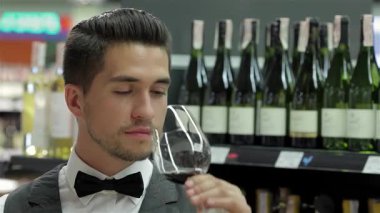 Zarif bir smokin giymiş dengeli bir erkek şarap garsonu, ahşap bir masanın üzerine eğilip zengin bir kırmızı şarabın kokusunun tadını çıkarıyor. Arkaplanda zarif bir şarap rafı, zarif şişeler sergileniyor, sofistike atmosferi...