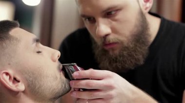 Şık bir berber dükkanında, odaklanmış bir berber müşterisinin sakalını titizlikle tımar eder, hassas ve sanatsal özelliklerini sergiler. Mahrem atmosfer, modern erkeklik ve özgüvenin altını çizerek, berberle yakışıklı adam arasındaki bağın altını çiziyor.