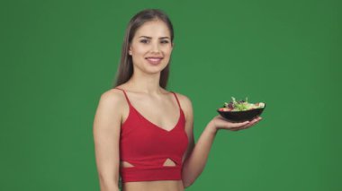 Renkli bir kase taze salata tutarak sağlıklı yaşama bağlılığını gösteren neşeli fiziği olan ışıl ışıl genç bir kadın. Canlı yeşil bir arka plan, gülüşü ve salatanın canlı renkleri duyguları çağrıştırıyor.