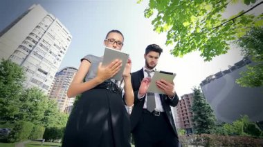 Kendinden emin bir işadamı ve başarılı bir iş kadını, modern mimari ve yemyeşil alanlarla çevrili canlı bir şehir parkında tabletleri üzerinde işbirliği yapıyorlar. Odaklanmış ifadeleri ve profesyonel kıyafetleri yeniliğe olan bağlılıklarını yansıtıyor.