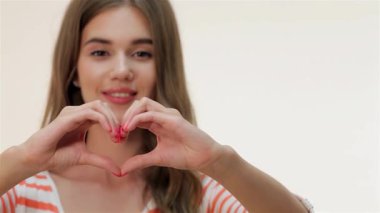 Neşeli kahverengi saçlı beyaz bir kız elleriyle bir kalp şekli oluşturur. Yumuşak beyaz bir arka plana neşe ve sıcaklık saçar. Parlak gülüşü ve neşeli jesti, sevgi ve sevgiyi ifade etmek için mükemmel bir ortam yaratıyor.