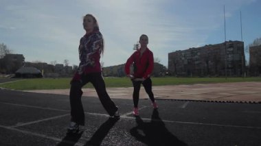 İki genç kız, yaklaşan bir yarış için bacak kaslarını hazırlayarak, güneşli pistte enerjik ısınma egzersizleri yapıyorlar. Enerjik atletik kıyafetleri ve odaklanmış ifadeleri bu koşucuların bağlılığını ve gücünü vurguluyor.