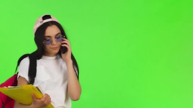 Canlı genç bir kadın, pozitiflik saçıyor, akıllı telefonuyla sohbet ederken parlak bir şekilde gülümsüyor, ders kitaplarını dengeliyor ve şık bir şapka takıyor. Canlı bir kromaanahtar arka planda, bu sahne gençlik enerjisinin ve akademik enerjinin özünü yakalar