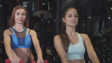 İki muhteşem genç kadın modern bir spor salonunda canlı kırmızı dambıllar kaldırıyor, güçlerini ve dostluklarını sergiliyorlar. Enerjik ifadeleri pozitiflik ve motivasyon yayıyor, sağlıklı bir yaşam tarzını ve spor zevkini somutlaştırıyor.